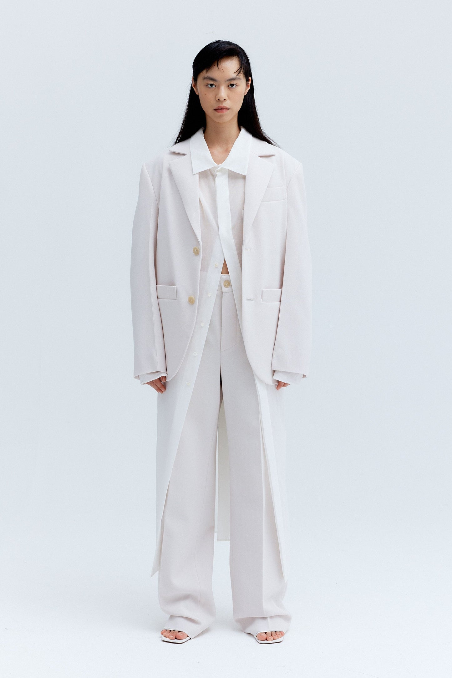 Cara standing in a studio wearing a straight pleat pants outfit in ice color, consisting of unisex wool blazer and woollen straight pleat pants