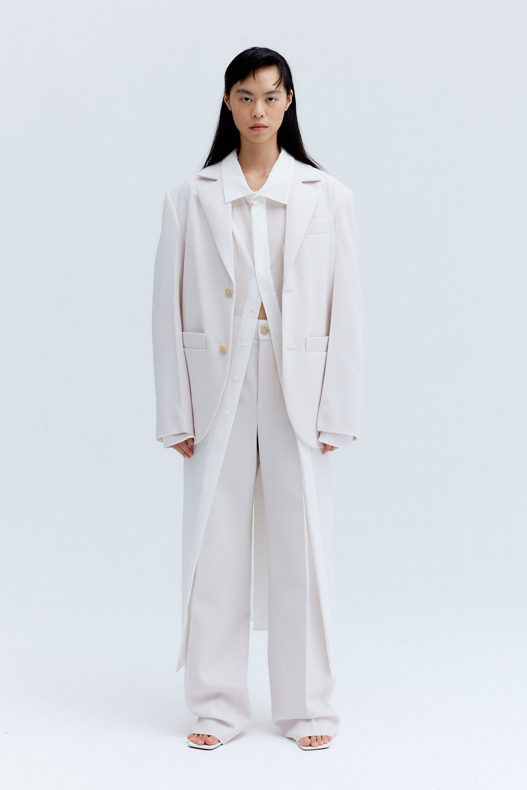 Cara standing in a studio wearing a straight pleat pants outfit in ice color, consisting of unisex wool blazer and woollen straight pleat pants