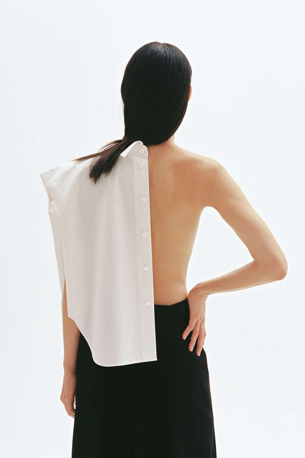 Model wearing asymmetric white cotton shirt with exposed back and button-down shoulder design