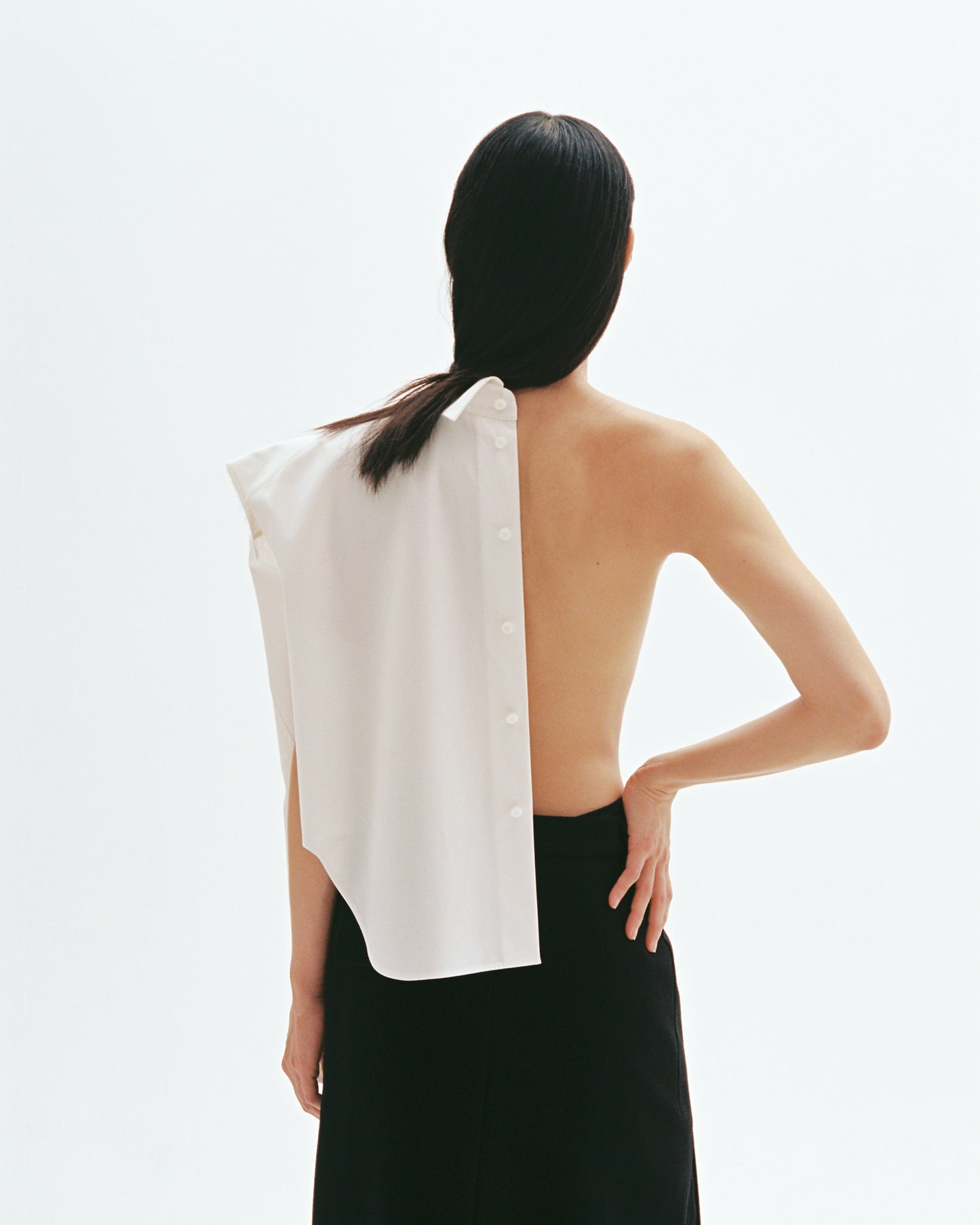 Model wearing asymmetric white cotton shirt with exposed back and button-down shoulder design