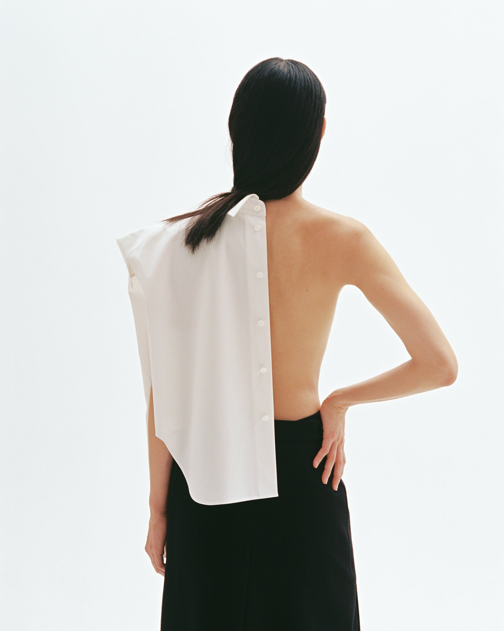 Model wearing asymmetric white cotton shirt with exposed back and button-down shoulder design