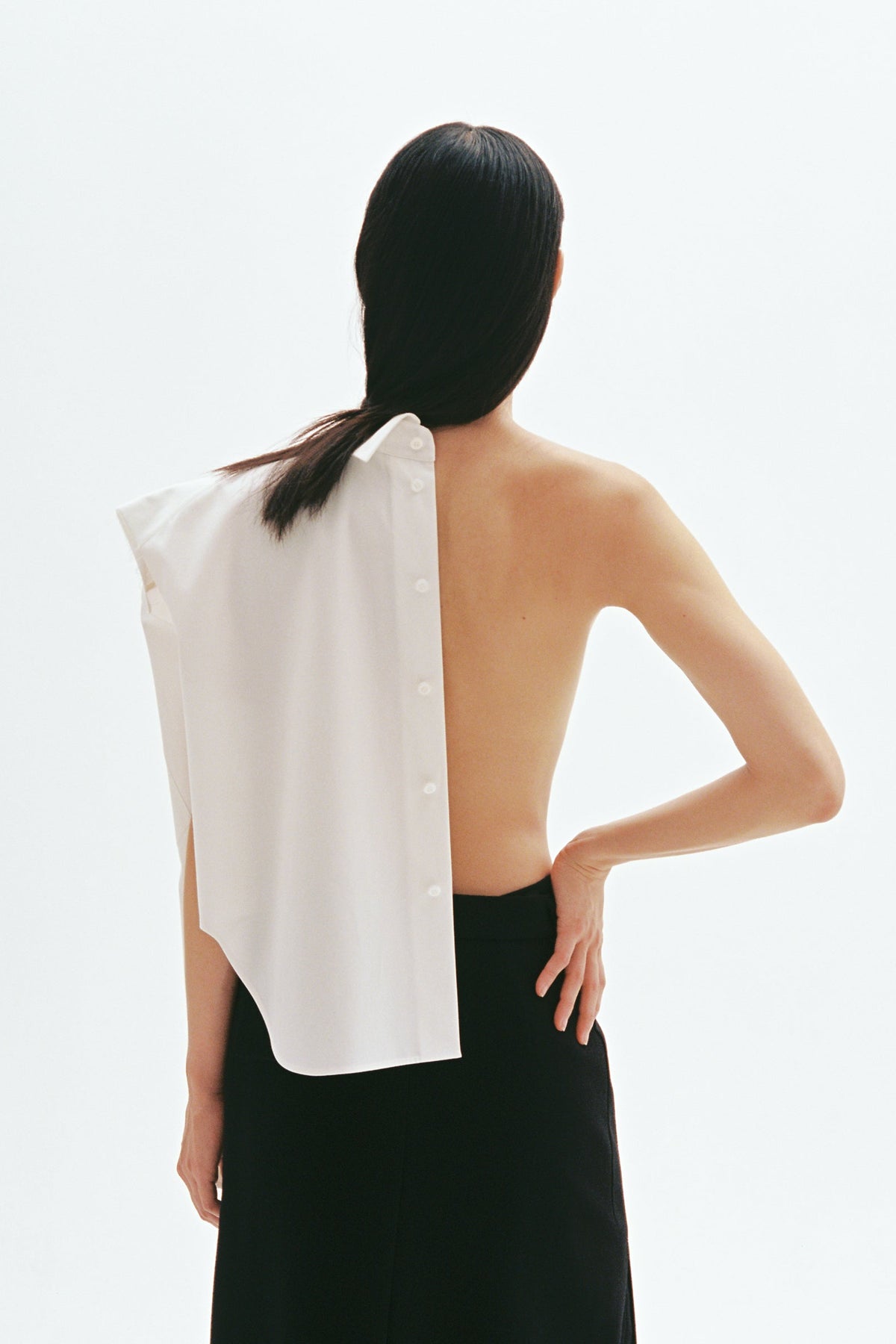 Model wearing asymmetric white cotton shirt with exposed back and button-down shoulder design